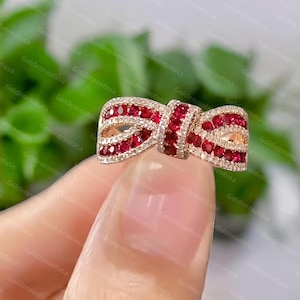 May include: A bow-shaped ring crafted from rose-toned metal, featuring a central knot and two loops encrusted with numerous small, vibrant red gemstones. The edges of the bow are lined with tiny, clear, sparkling stones, creating a detailed design.
