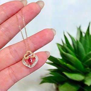 May include: A rose gold necklace featuring a pendant with a central oval red gemstone, surrounded by a decorative frame set with small clear stones, suspended from a delicate rose gold chain with a fine link design.