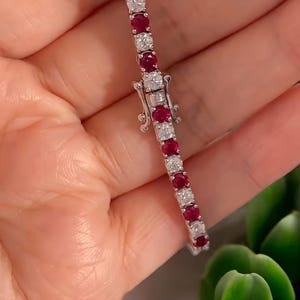May include: A tennis bracelet featuring alternating round, vibrant red rubies and sparkling white diamonds. The bracelet is set in a silver metal and has a secure clasp. The jewelry is displayed against a soft background.