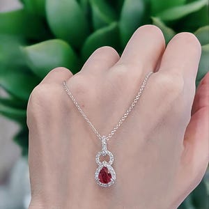 May include: A silver necklace with a pendant featuring a pear-shaped, deep red gemstone surrounded by a halo of small, clear gemstones. The pendant is suspended from a delicate chain. The background is a blurred green plant.