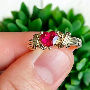May include: A gold ring featuring a heart-shaped, faceted red gemstone. The gemstone is set in a gold band with an X and O design. The ring is shown against a blurred green background.