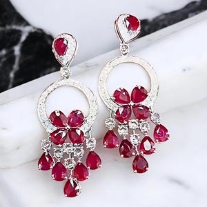 Peut inclure: Une paire de boucles d'oreilles pendantes élégantes avec un motif floral. Elles présentent des pierres précieuses rouges vibrantes et des accents blancs étincelants, serties dans un métal argenté. Forme de goutte.