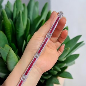 Könnte beinhalten: Ein silbernes Armband mit einer Reihe oval geschliffener, leuchtend roter Edelsteine. Das Armband ist mit klaren, funkelnden Steinen und dekorativen silbernen Gliedern verziert. Der Schmuck wird vor einem Hintergrund aus grünem Laubwerk präsentiert.