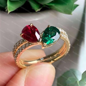 May include: A gold ring featuring two pear-cut gemstones, one deep red and one vibrant green, set side-by-side. The band is split into two thin, polished gold shanks, each encrusted with a row of small, clear, round-cut white stones.