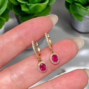 May include: A pair of gold hoop earrings with a ruby-red oval gemstone center, surrounded by a halo of small, clear stones. The earrings are held up by a person's fingers, with green plants in the background.