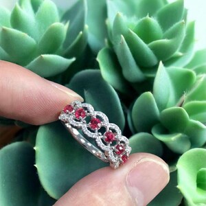 Könnte beinhalten: Ein silberner Ring mit einem sich wiederholenden Muster aus roten Edelsteinen und kleinen weißen Diamanten. Der Ring wird vor dem Hintergrund grüner Sukkulenten präsentiert, was das Design und den Farbkontrast des Schmucks hervorhebt.