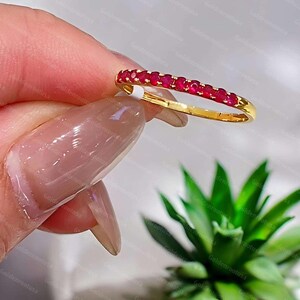 Puede incluir: Un anillo de banda fina de oro con una hilera de pequeños rubíes redondos de color rojo vibrante engastados a lo largo de la mitad superior. El metal tiene un acabado pulido y las piedras preciosas están dispuestas en una línea continua.