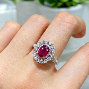 Puede incluir: Un anillo de tono plateado con una piedra preciosa de color rojo intenso de corte ovalado en el centro, rodeada por un halo de pequeñas piedras claras de corte redondo. La banda está dividida y adornada con piedras claras adicionales.