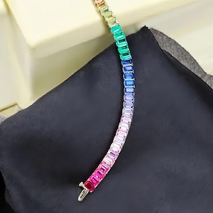Könnte beinhalten: Ein farbenfrohes Armband mit rechteckigen Edelsteinen in Regenbogenfarben, darunter Rosa, Rot, Orange, Gelb, Grün, Blau und Lila. Das Armband ist auf silberfarbenem Metall gefasst und liegt auf einer schwarzen Oberfläche.