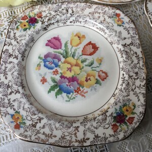 Puede incluir: Un plato cuadrado de cerámica de color crema con un borde dorado y un diseño floral. El centro del plato presenta un diseño de punto de cruz de tulipanes coloridos y otras flores.