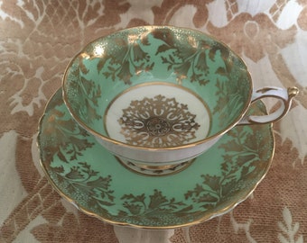Puede incluir: Un juego de taza y platillo de té verde y dorado con un patrón floral. La taza y el platillo tienen un borde festoneado y están decorados con ribetes dorados.