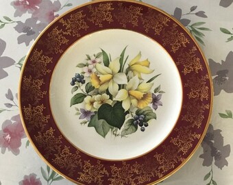 May include: A white ceramic plate with a gold rim and a burgundy border. The plate features a floral design with yellow daffodils, green leaves, and purple berries.
