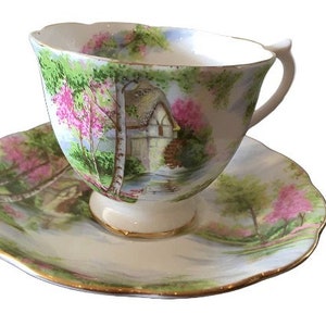 Puede incluir: Una taza de té y un platillo blancos con un patrón floral. El patrón presenta flores rosas, hojas verdes y una pequeña cabaña.