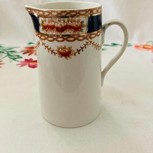 May include: A white ceramic milk jug with a gold and blue floral design. The jug has a handle and a rounded base.