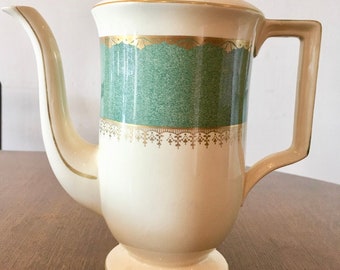 May include: A white ceramic coffee pot with a green and gold design. The pot has a lid with a handle and a spout. The base of the pot is round and has a gold trim.