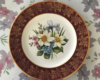 May include: A white ceramic plate with a gold and burgundy rim. The plate features a floral design with white, yellow, and blue flowers.