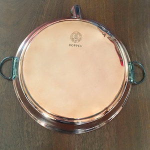 Puede incluir: Una bandeja redonda de servir de cobre con dos asas. La bandeja tiene un borde elevado y una superficie lisa. La bandeja está grabada con el texto "Registered Trade Mark" y "COPPEK".