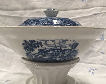 Op de afbeelding: Een witte keramische serveerschaal met een blauw-wit landschapsontwerp, inclusief deksel en een kleine knop. De schaal staat op een witte sokkel. Het ontwerp van de schaal toont bomen, gebouwen en een landelijke scène.