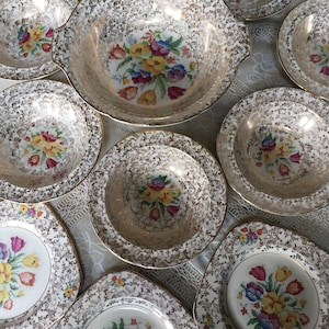 May include: A set of vintage floral patterned plates and bowls with gold trim. The plates and bowls have a white background with a gold floral pattern and a colourful floral design in the centre.