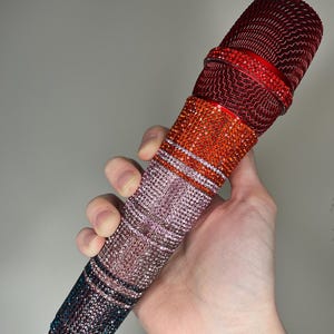 May include: A microphone adorned with rhinestones, featuring a colour gradient. The top is deep red, fading to orange, pink, and dark blue at the base. The microphone is held in a hand.