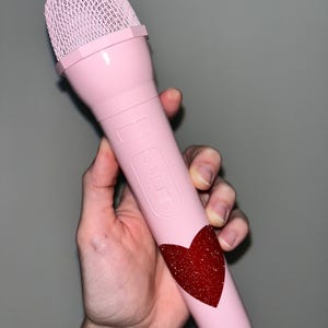 May include: A pink microphone with a black button and a red glitter heart design. The microphone has a black mesh grille.