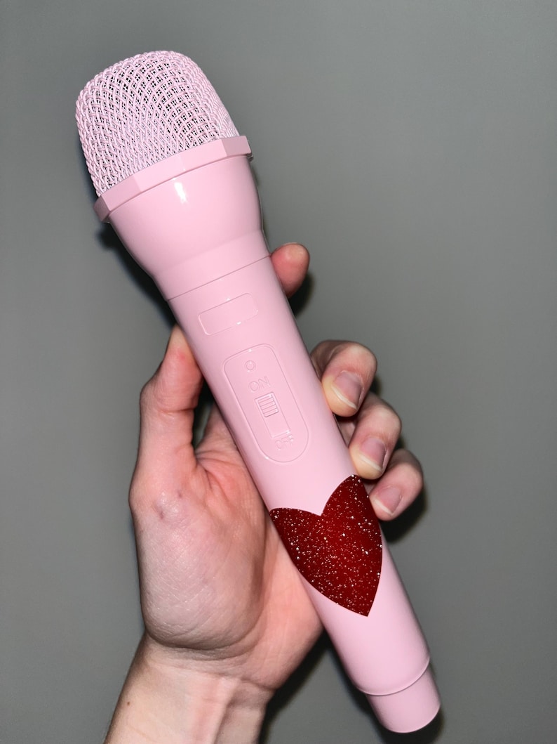 May include: A pink microphone with a black button and a red glitter heart design. The microphone has a black mesh grille.