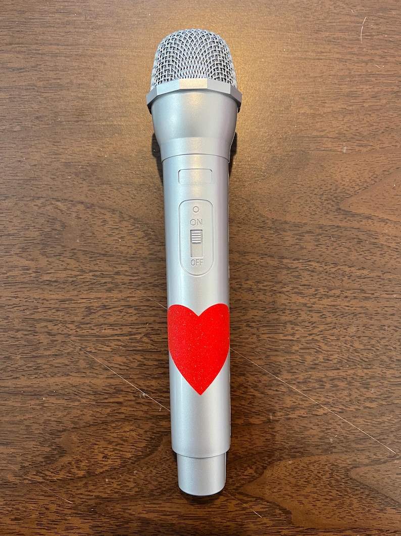 May include: A silver microphone with a red heart sticker on the body. The microphone has an on/off switch.