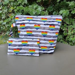 May include: Two zippered pouches with a rainbow heart pattern on a white and gray striped background. The larger pouch has a handle.