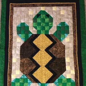 May include: A green and brown patchwork quilt with a turtle design. The turtle's shell is brown with a yellow diamond pattern. The turtle's head and legs are green.