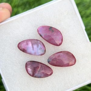 May include: Four pink and purple gemstone cabochons in a white box. The cabochons are oval shaped and have a smooth, polished surface.