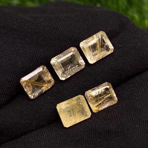 Pode incluir: Cinco pedras preciosas de quartzo rutilado douradas em forma retangular. As pedras preciosas são translúcidas e têm um brilho dourado.
