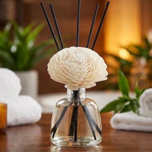 May include: A clear glass diffuser bottle with a cream-coloured, flower-shaped top and black reeds. The bottle sits on a wooden surface, with greenery and white towels in the background. The diffuser is designed for home fragrance.