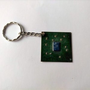 Peut inclure: Un porte-clés en forme de circuit imprimé vert avec un anneau et une chaîne argentés. Le circuit imprimé a un petit carré bleu au centre.