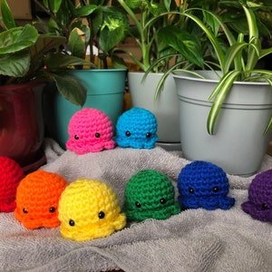 May include: A collection of nine crocheted octopus toys in a rainbow of colors: red, orange, yellow, green, blue, purple, pink, and teal. Each octopus has small black eyes. The toys are displayed on a gray towel with potted plants in the background.