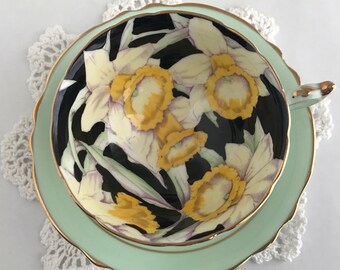 May include: A vintage teacup and saucer set with a black background and a floral pattern of yellow and white daffodils. The saucer is a pale green color with a gold trim.