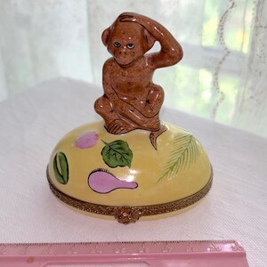 May include: A decorative trinket box with a brown monkey figurine on top. The box is yellow with painted floral and leaf designs. The box has a gold-colored metal trim and a pink ruler is in the foreground.