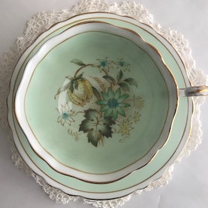 May include: A vintage teacup and saucer set with a delicate floral design. The cup and saucer are a pale green color with a gold trim. The floral design features yellow, green, and blue flowers.