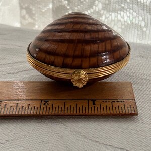 May include: A decorative clam shell-shaped trinket box. The box is brown with a wood grain pattern and a gold-colored rim. The box is approximately 2 inches long.