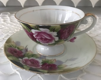 May include: A white teacup and saucer set with a floral rose design in shades of pink, green, and gold. The teacup has a gold rim and handle, and the saucer has a matching design. The set is on a white surface.