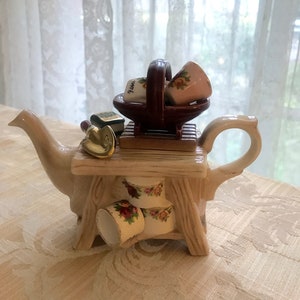 May include: A decorative teapot with a whimsical design. The teapot is beige with a wooden table-like structure on top, holding a basket with miniature cups. Two cups are placed below the table. A small gold bell and a miniature book are also on the table.