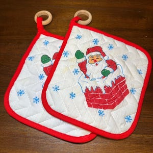 May include: Two quilted Christmas pot holders with a red trim and wooden hanging rings. Each pot holder features a Santa Claus design emerging from a red chimney, surrounded by blue snowflakes on a white background.