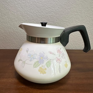 Puede incluir: Tetera de cerámica blanca con asa y tapa negras. La tetera está decorada con un estampado floral en colores pastel, incluyendo azul, rosa y amarillo. Una banda plateada rodea el centro de la tetera.