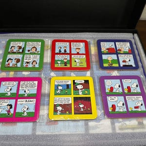 Peut inclure: Ensemble de six dessous de verre carrés avec des bandes dessinées de la série Peanuts. Chaque dessous de verre a une bordure de couleur différente, notamment verte, rouge, bleue, rose, jaune et violette. Les dessous de verre sont emballés dans une boîte noire.