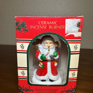 May include: Ceramic incense burner in the shape of Santa Claus, packaged in a red box with green accents. The box reads "CERAMIC INCENSE BURNER" and "WITH 2 POTPOURRI INCENSE CONES". Santa is wearing a red suit with white trim and green gloves.