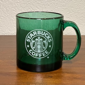 Puede incluir: Taza de café de vidrio verde con el logotipo de Starbucks en blanco. La taza tiene una base negra y un asa verde. El logotipo de Starbucks presenta una sirena blanca con las palabras "Starbucks Coffee" en un círculo blanco.
