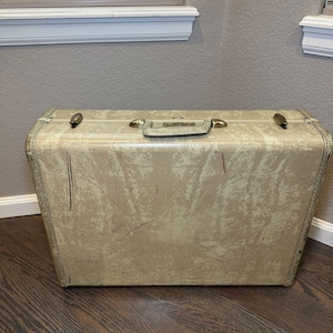Könnte beinhalten: Ein beigefarbener Vintage-Koffer mit messingfarbener Hardware, einschließlich Griff und Verschlüssen. Der Koffer weist Gebrauchsspuren auf, mit Kratzern und einem abgenutzten Finish. Der rechteckige Koffer steht vor einem neutralen Hintergrund.