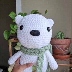 May include: White crocheted polar bear plush toy wearing a green scarf. The bear has black eyes and a black nose.