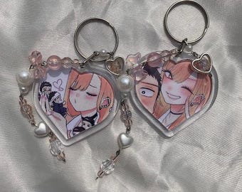 以下が含まれることがあります： アニメスタイルのイラストが描かれた、ハート型のキーホルダー2つ。各キーホルダーには、シルバーのリング、ハート型のチャーム、パールとピンクのビーズが付いたビーズアクセントが付いています。イラストは、ピンクの髪と笑顔の顔をしたキャラクターを描いています。