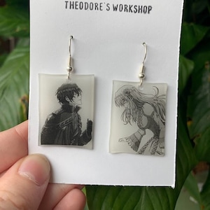 Puede incluir: Un par de pendientes colgantes con paneles rectangulares de vidrio transparente. Los paneles presentan imágenes en blanco y negro de personajes de anime. La tarjeta detrás de los pendientes dice "Theodore's Workshop".