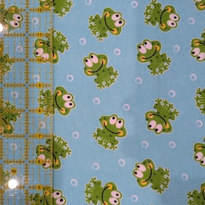 May include: Light blue fabric with a repeating pattern of green cartoon frogs and small white circles. The frogs have white eyes, yellow smiles, and are in various poses. The fabric is likely cotton or a similar material.